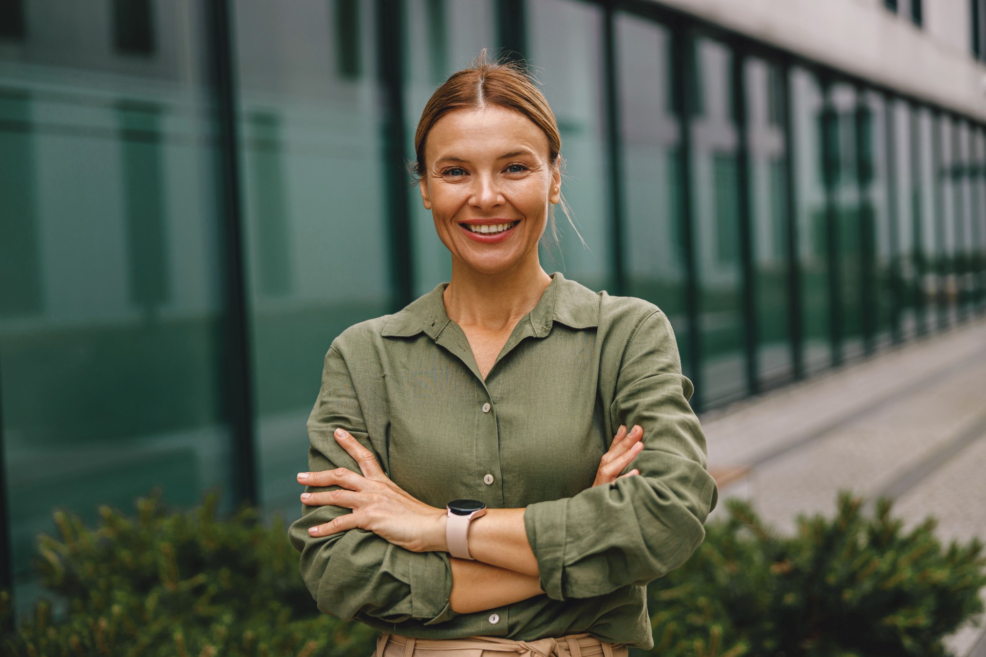 Woman entrepreneur with crossed hands is standing on office building background and looking camera